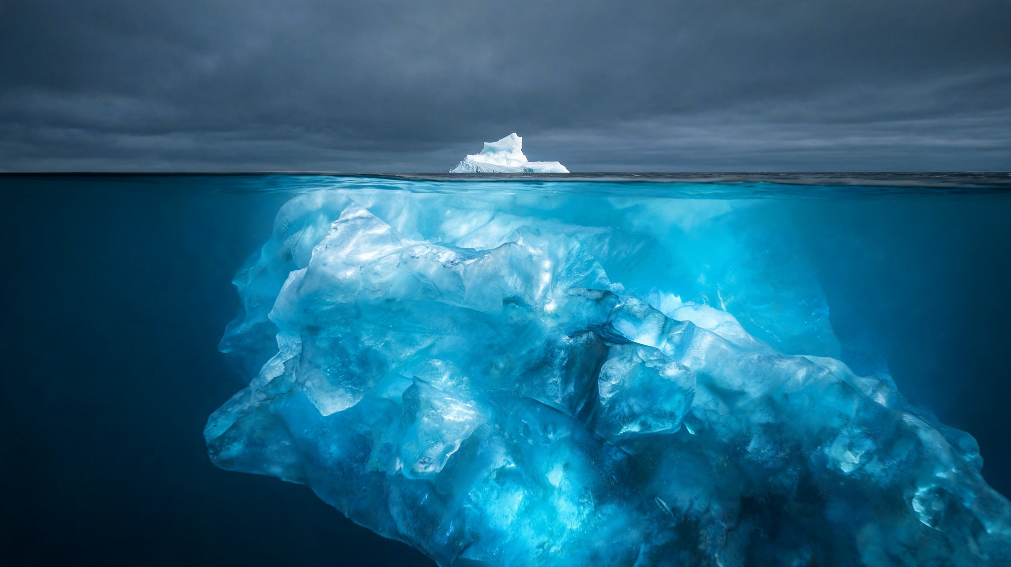 Dramatic split-level photo of an iceberg with a small visible tip above water and a vast glowing mass below the surface, illustrating the hidden total cost beneath a low sticker price.